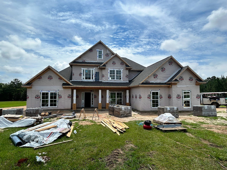 Partially built house with exposed framing, surrounded by stacks of lumber and construction materials on grassy yard, cloudy sky overhead, white bus parked nearby