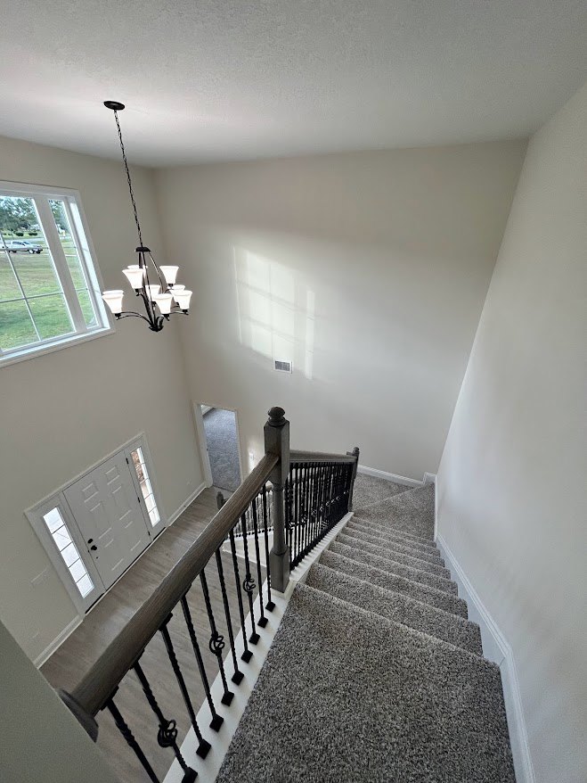 Staircase with matte black railing, modern chandelier overhead, white walls, and large window revealing parked car outside