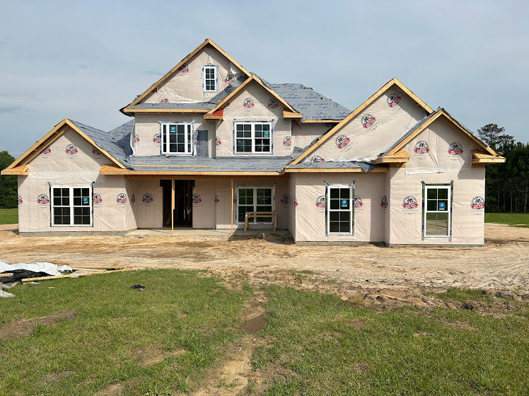 Modern cottage-style home under construction with exposed framing, large windows, and lush green grass in the yard; wooden bench placed near the unfinished exterior.
