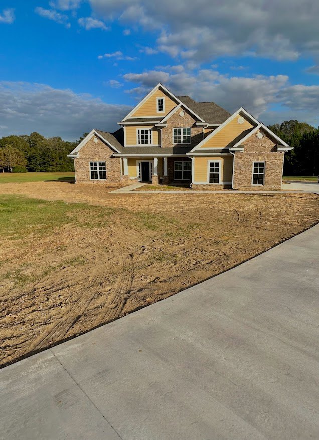 Modern custom home with large front door, paved driveway, manicured lawn, and surrounding trees under a blue sky with scattered clouds
