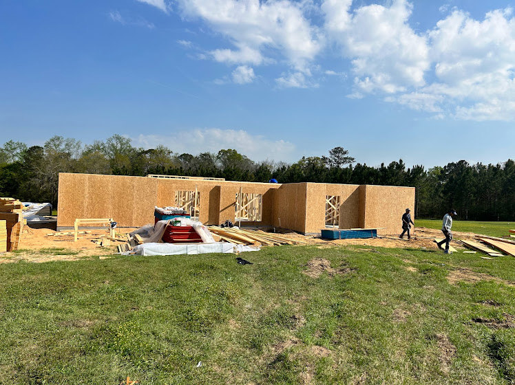 Modern home under construction with exposed framing, workers walking on grassy lot, surrounded by trees under partly cloudy sky
