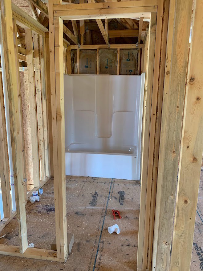 Unfinished bathroom with exposed wood planks, white shower stall featuring a hole in the wall, brown bag with blue writing, white plastic pipe on plywood floor