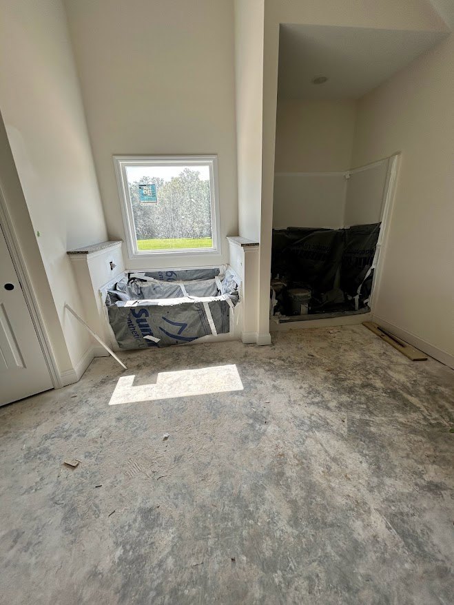 Bathroom with freestanding white tub beneath a large window, unfinished plaster walls, construction materials on dusty floor, exposed wall opening.