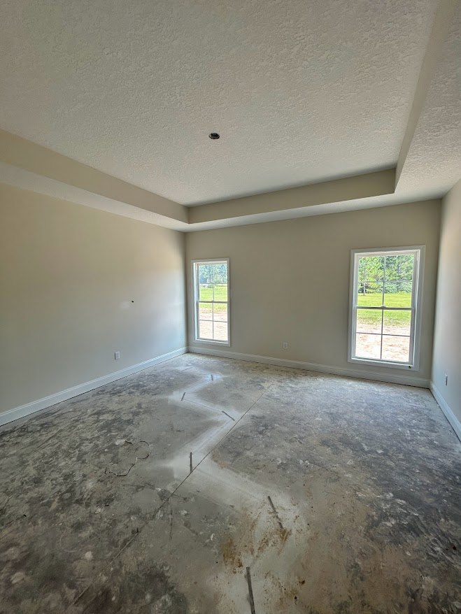 Concrete floor with circular holes, large windows overlooking grassy field, white plaster ceiling featuring a central opening, minimalist interior with natural daylight.