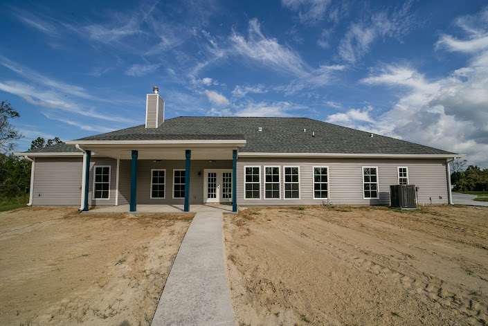 Two-story house with white siding and dark roof, large windows, surrounded by trees, dirt path leading to front entrance, cloudy sky overhead