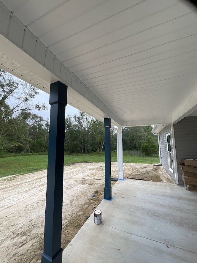 Covered porch with blue pillars, white ceiling, and concrete floor overlooking a dirt road, surrounded by grass and trees.