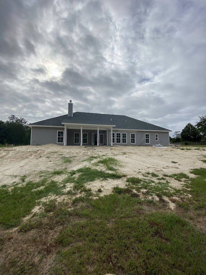 Partially built house with white roof, exposed framing, front porch, surrounded by dirt field and scattered trees under cloudy sky