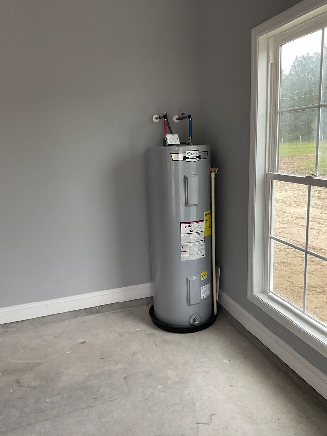 Large grey water heater with yellow label, surrounded by pipes, positioned on concrete floor near white-framed window overlooking grassy field