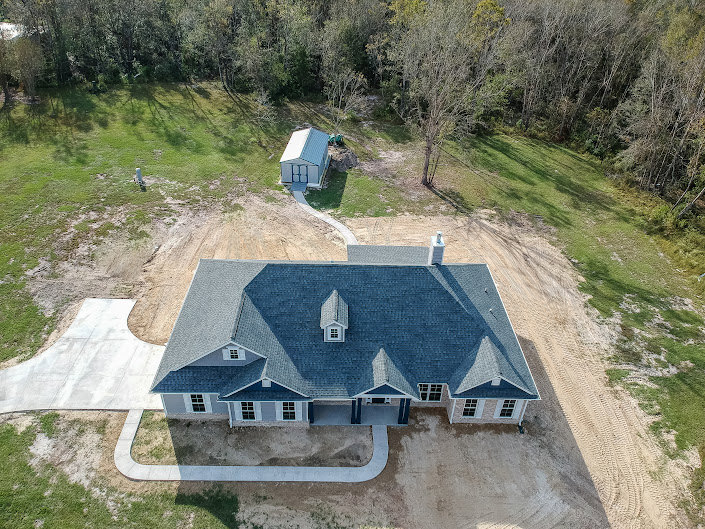 Two-story house with light siding, attached garage, paved driveway, manicured lawn, mature trees, and shrubs bordering the property