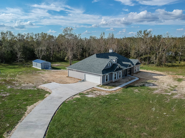 Two-story house with light siding, shingle roof, paved driveway, manicured lawn, and mature trees in the background under a partly cloudy sky