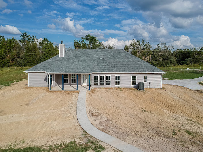 Two-story cottage with white siding and dark roof, surrounded by grass and trees, dirt path leading to front entrance under partly cloudy sky