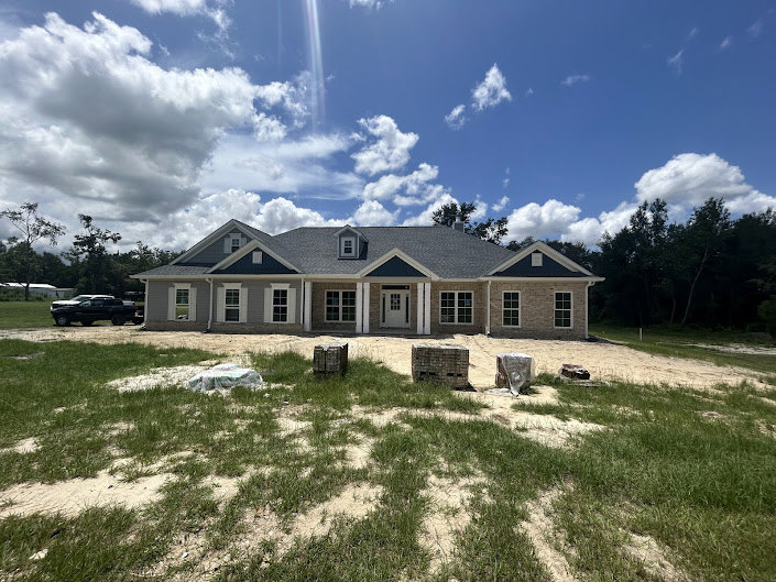 Two-story home with white siding and large windows, surrounded by a spacious green lawn, mature trees, and under a bright blue sky with scattered clouds