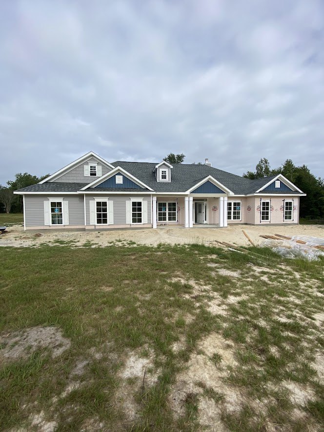 Two-story home with blue roof, white siding, large front porch, expansive green lawn, and cloudy sky overhead