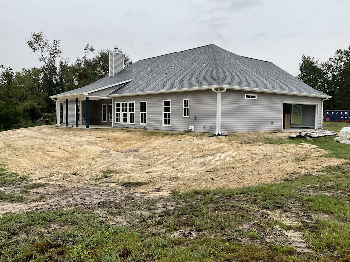 Single-story home with light siding, surrounded by a dirt yard, sparse grass, and mature trees under a clear sky