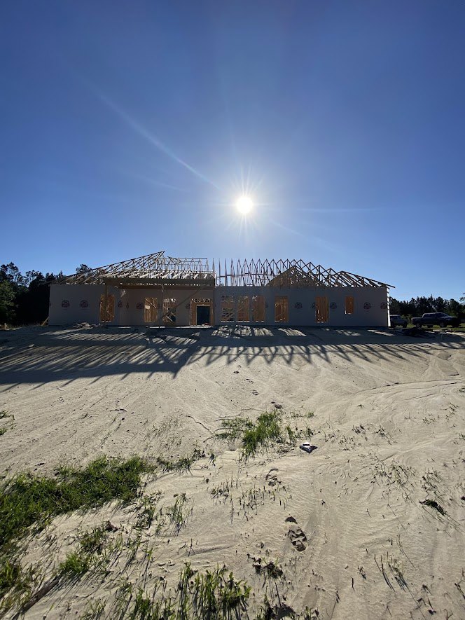 Wood-framed custom home under construction with exposed beams, ladder leaning against structure, sandy ground with patches of grass, wire fence, and bright sun in clear blue sky