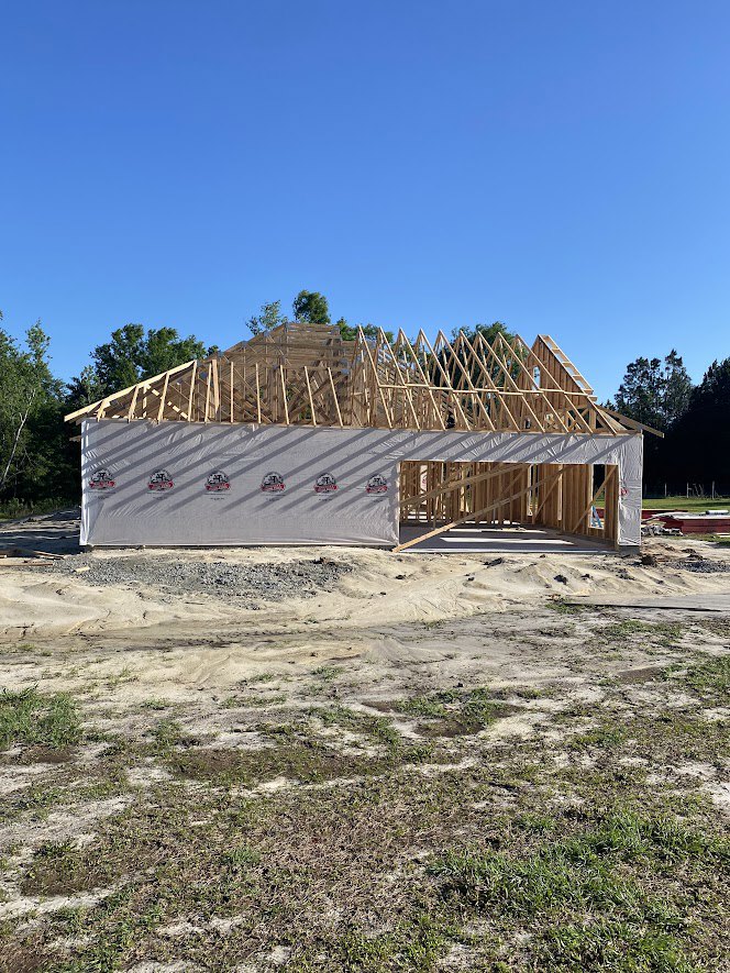 Partially built house framed with exposed lumber, white tarp covering sections, dirt road and grassy field in foreground, mature trees and blue sky in background