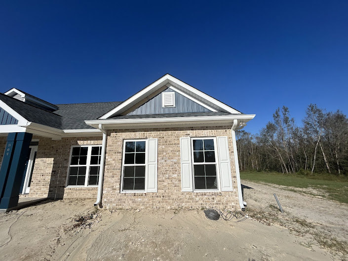 Two-story home with light siding, multiple windows, and gabled roof, set beside a dirt road with mature trees and open sky in the background