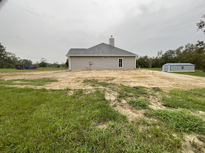 Single-story cottage with light siding, surrounded by a dirt yard, small shed to the side, grassy patches, mature trees, and open rural landscape under a partly cloudy sky