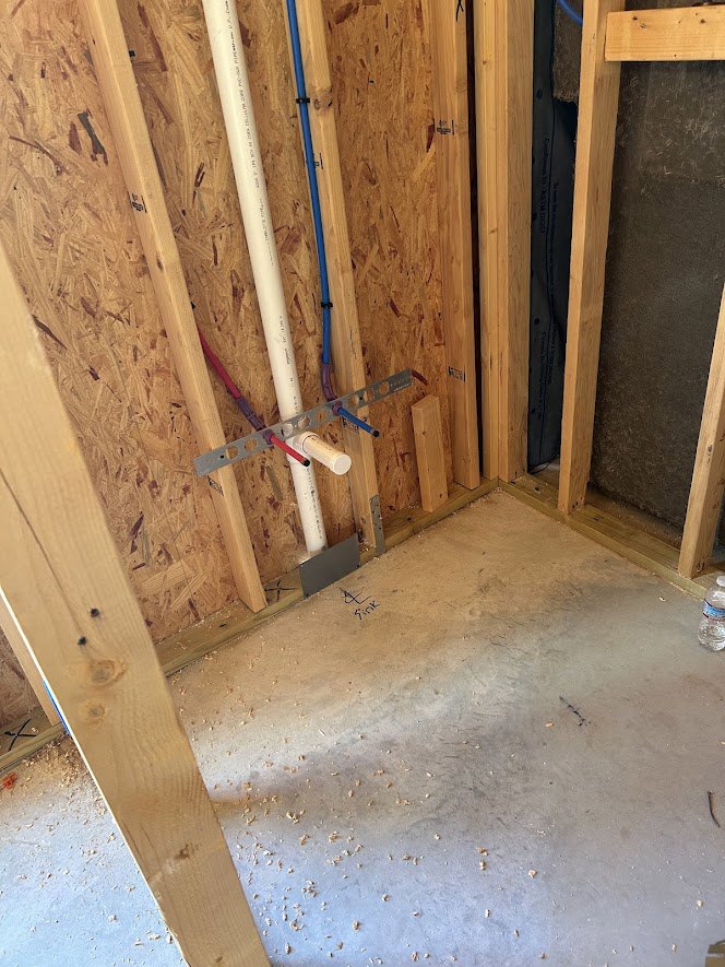 Exposed wooden framing with electrical wires and plumbing pipes, black insulation behind studs, plywood floor scattered with wood chips, blue pencil resting on wood surface
