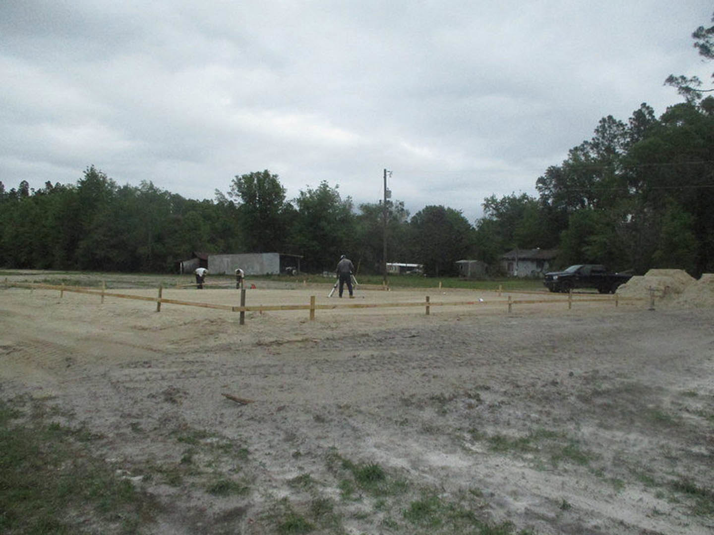 Brick custom home with white trim set on grassy lot, mature trees in background, cloudy sky overhead, group of people playing on lawn, parked car visible near road.