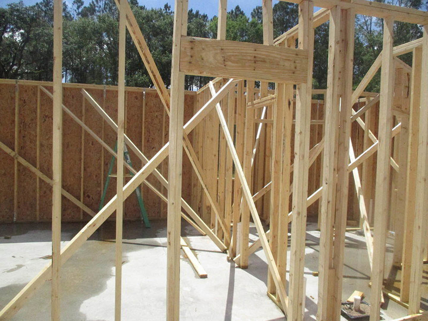 Wood-framed house under construction with exposed beams, concrete foundation, and ladder, surrounded by trees