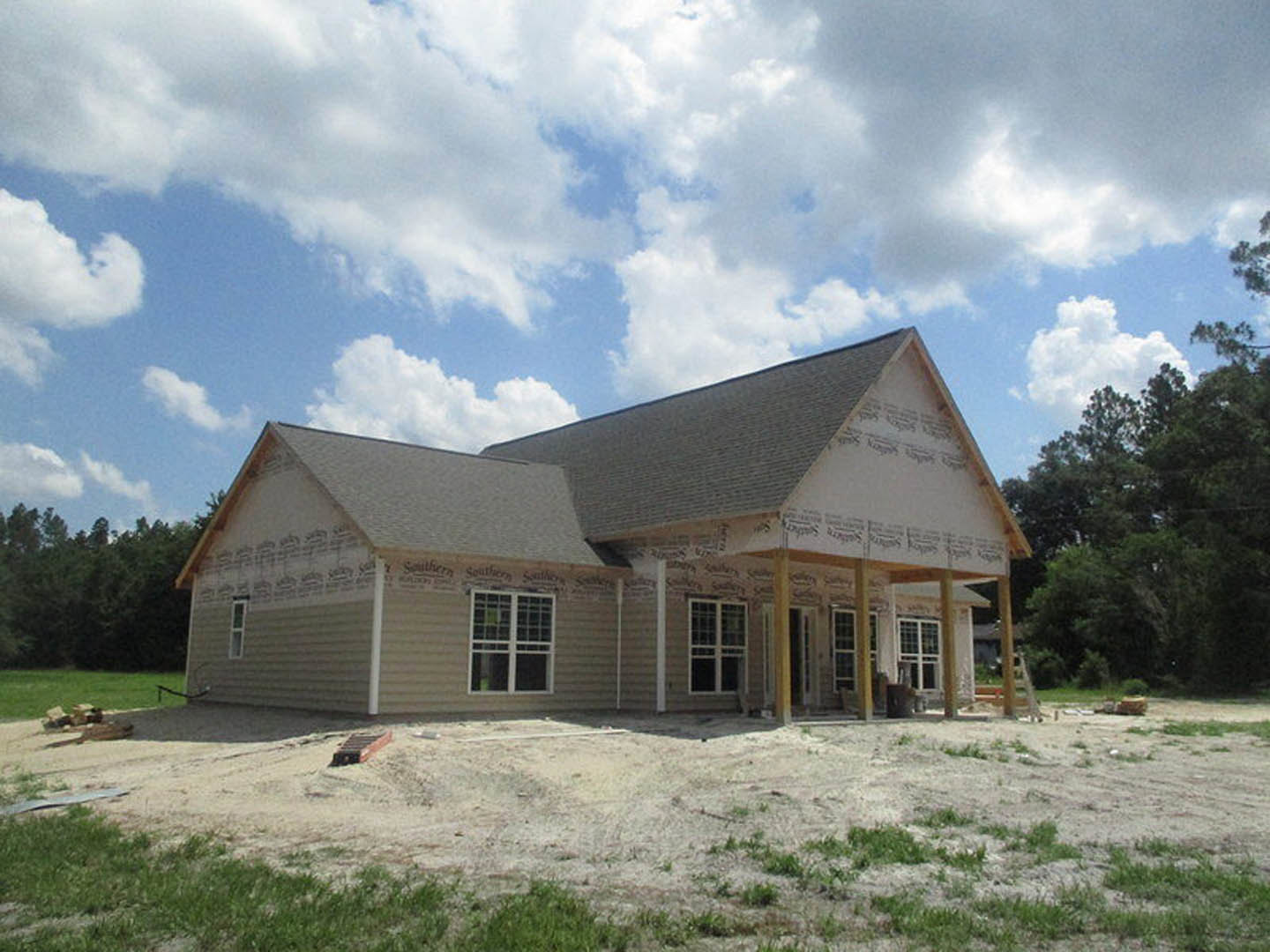 Partially built house with exposed framing, unfinished porch, and scattered trees on grassy lot under cloudy sky