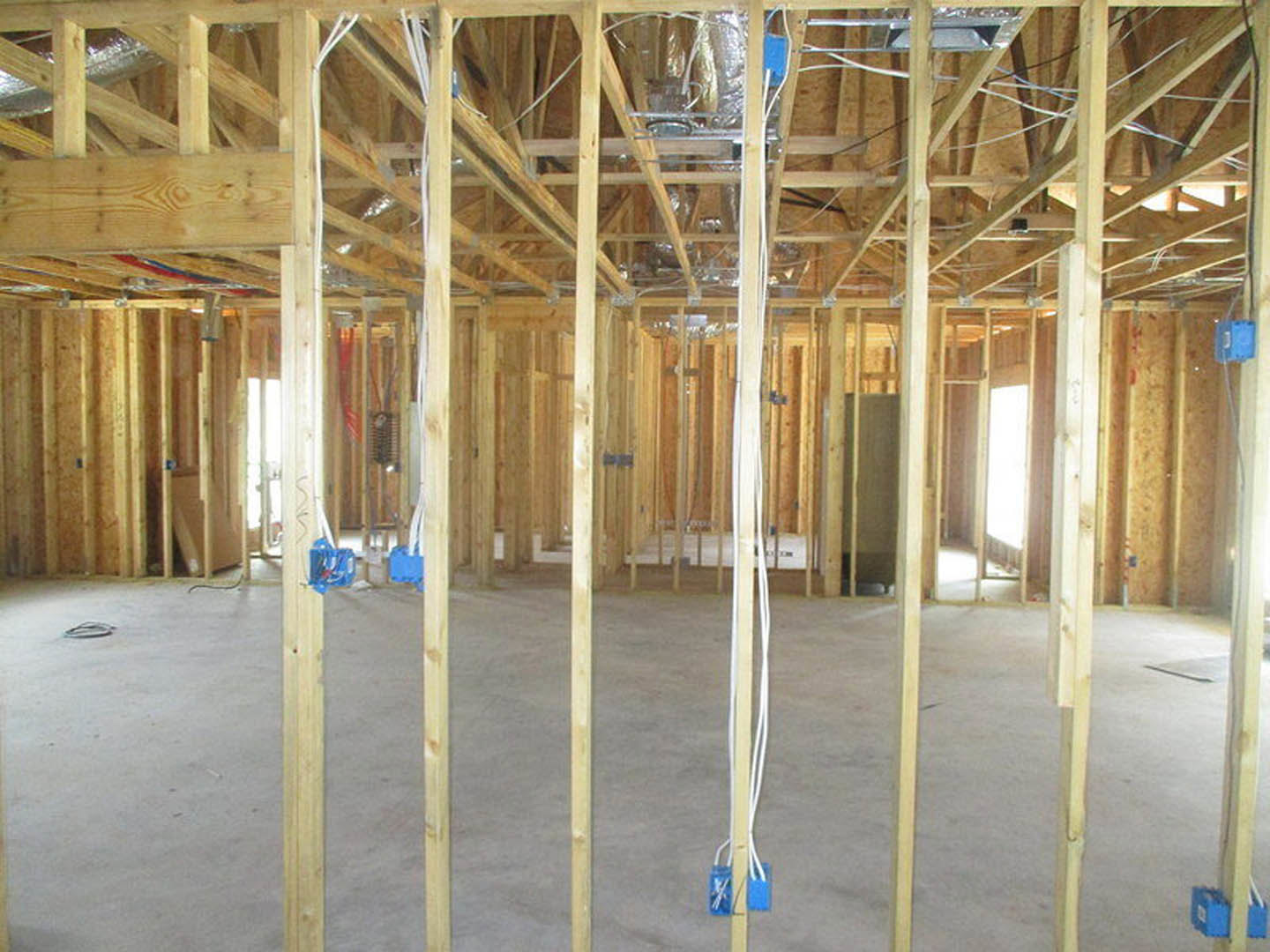 Wood-framed room under construction with exposed beams, blue electrical box mounted on wall, loose wires hanging, and building insulation visible in ceiling.