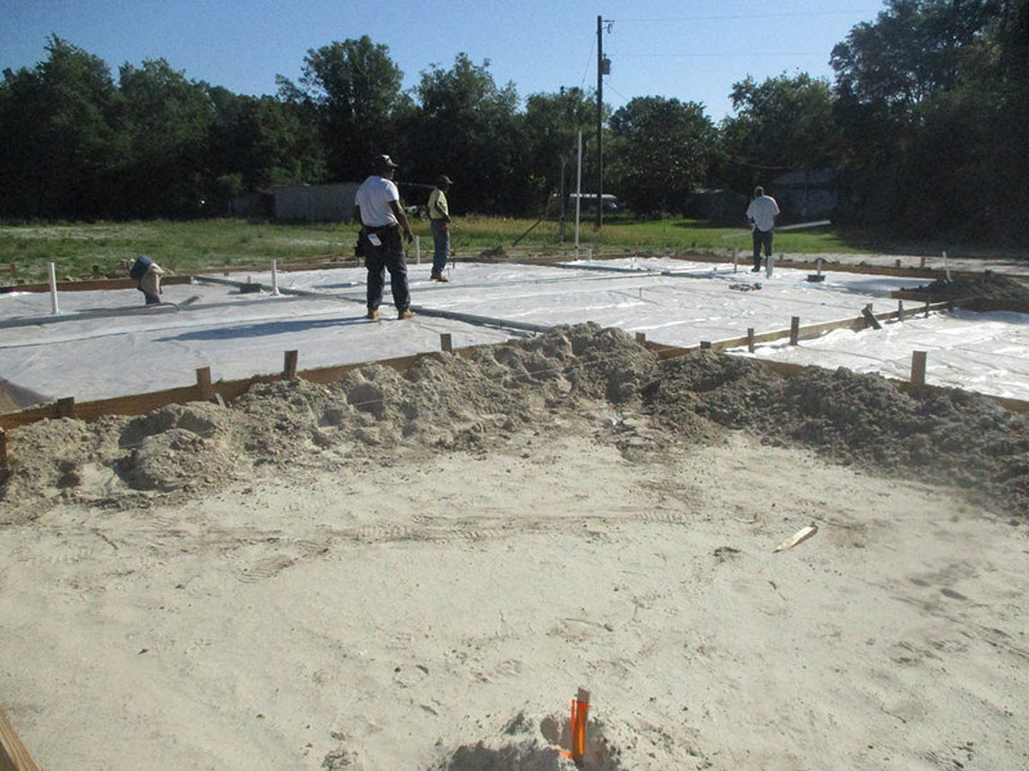 Framed foundation of custom home under construction with several workers on sandy soil, surrounded by trees and open sky
