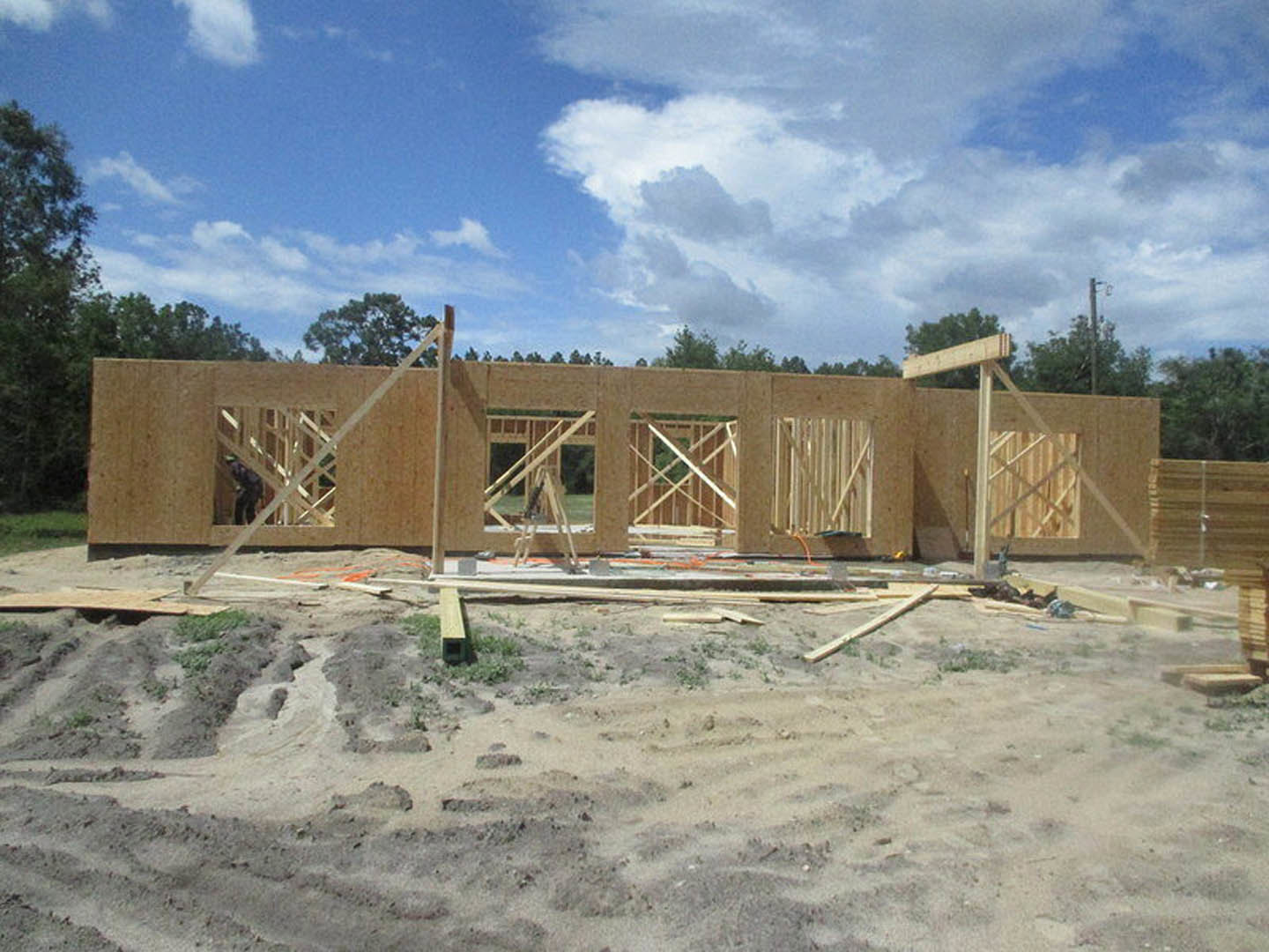 Framed house under construction with exposed wood beams, foundation, ladder, and workers on site beneath partly cloudy blue sky