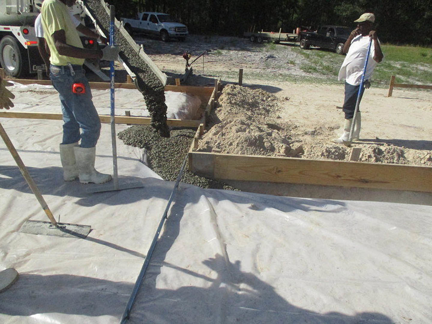 Men in work boots and jeans assembling foundation framing on a dirt construction site, white truck parked nearby, trees in background.