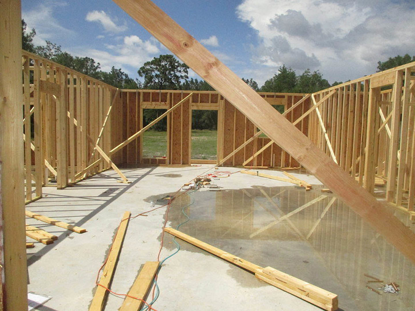 Wooden framing and beams set atop a wet concrete foundation, construction materials scattered, green field and trees in background under a cloudy blue sky