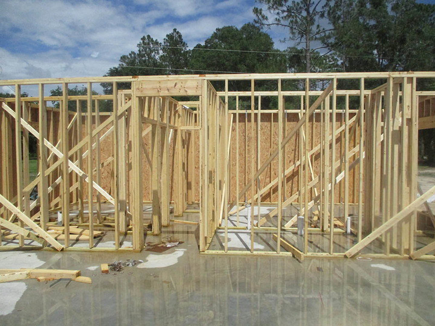Wooden house frame under construction with exposed lumber beams, wet concrete floor, and trees visible in the background.