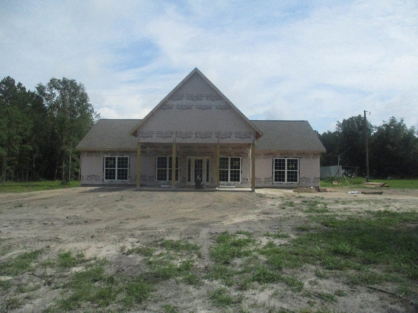 Partially built house with triangular roof, exposed framing, dirt lot in foreground, scattered grass, trees and blue sky with clouds in background