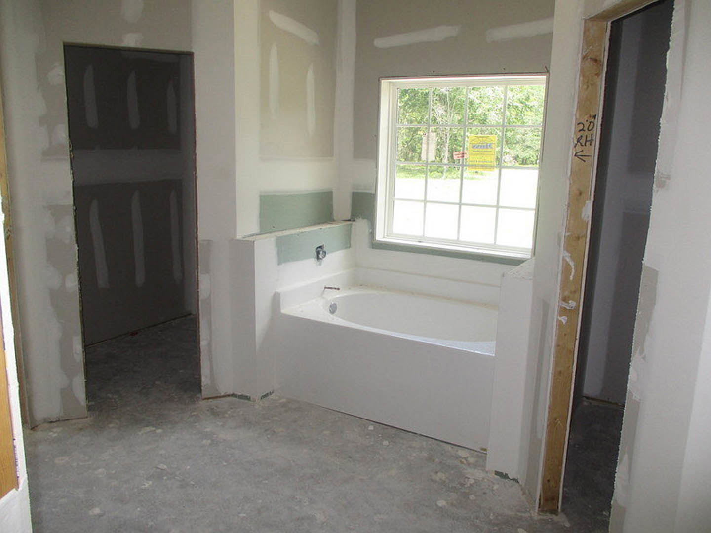 Bathroom under construction featuring a white bathtub, unfinished plaster walls, exposed plumbing, and a window with a yellow sign taped to the glass.