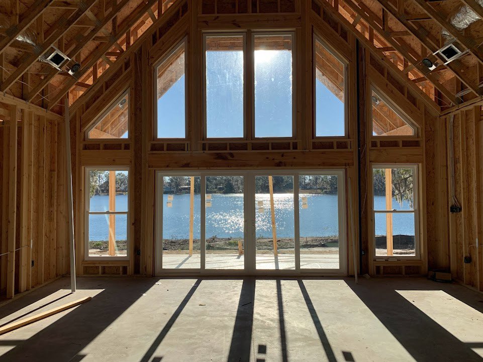 Open-concept room featuring large wood-framed windows overlooking water and trees, sunlight casting fence shadows across polished concrete floor, exposed wooden beams, and hardwood