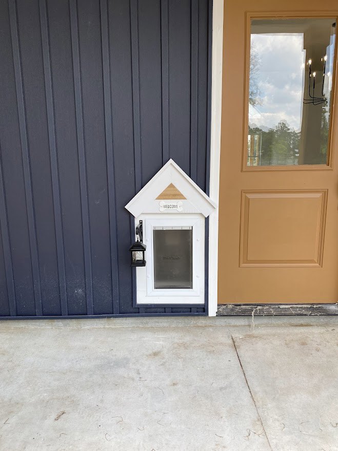 Small custom dog house with black door and window, light fixture above entry, concrete ground surface