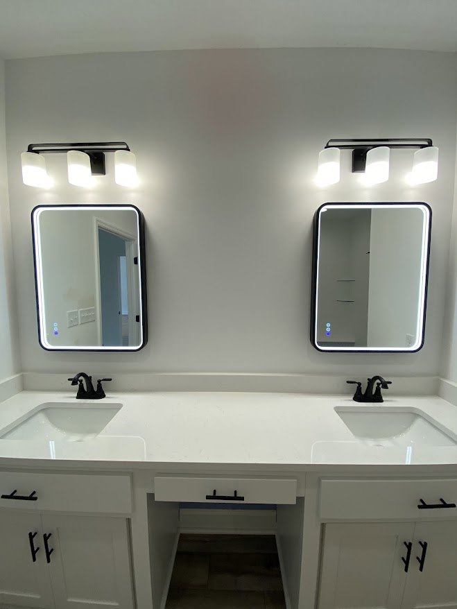 Double vanity bathroom with two white sinks, black faucets, large wall mirror, white door, and modern wall-mounted light fixture.