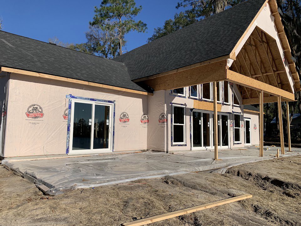 Wood-framed house under construction with exposed beams, white-framed window, wooden planks on ground, partially finished roof, and trees in background