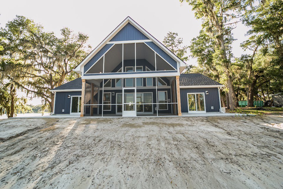 Two-story house with white-framed windows, covered porch, sandy dirt road leading to entrance, surrounded by trees under clear sky