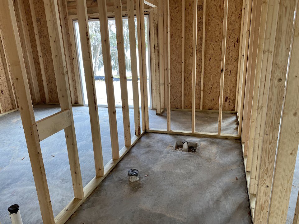 Concrete floor with cutout, exposed wood framing, white pipe with black cap, stacked lumber, large window on unfinished wall