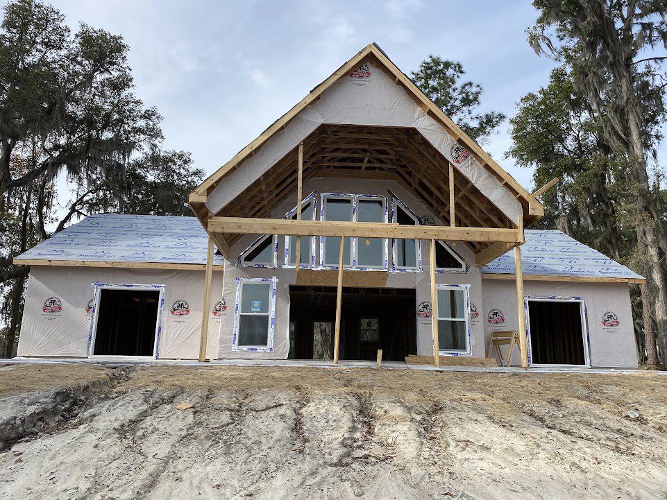 Wood-framed house under construction with exposed beams, white rectangular window frame, dirt foreground, and trees in the background
