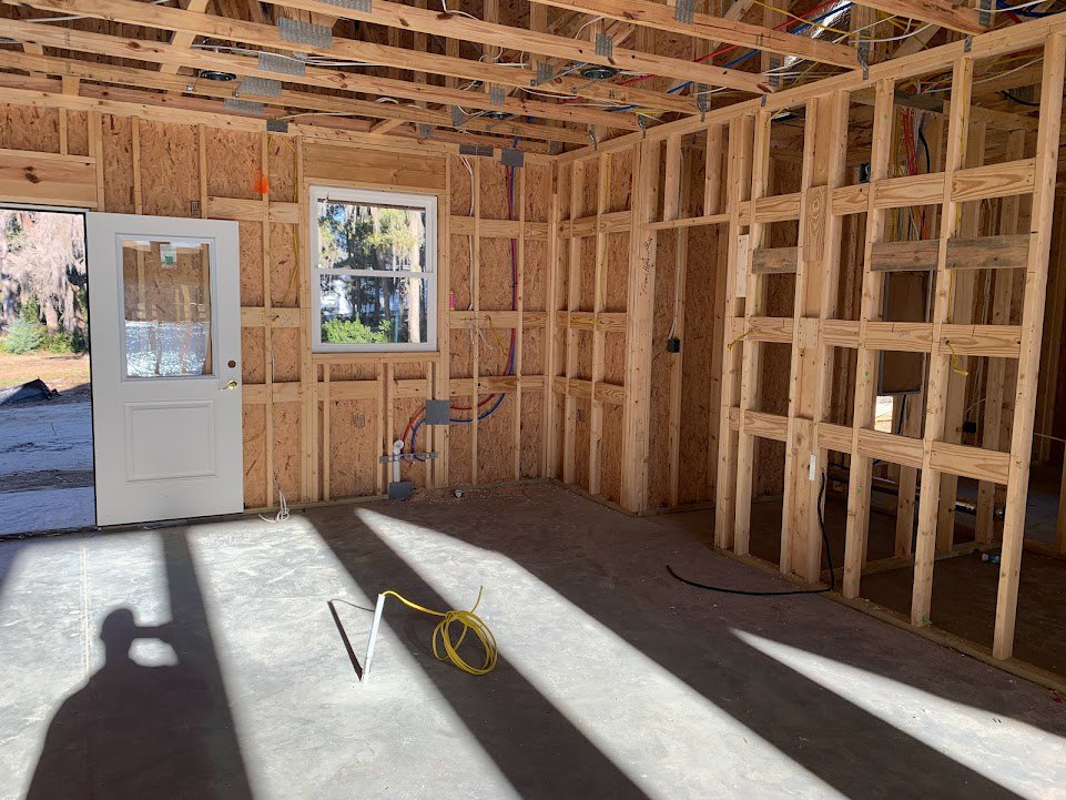 Unfinished room with exposed wood beams, white door with glass window, yellow electrical wires on floor and pole, window showing trees and snow outside