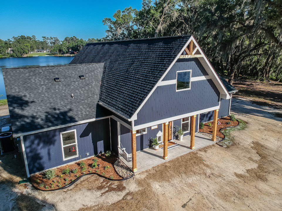 Two-story cottage with gray siding and white trim, surrounded by mature trees and landscaped garden beds, overlooking a calm lake in the background under a partly cloudy sky