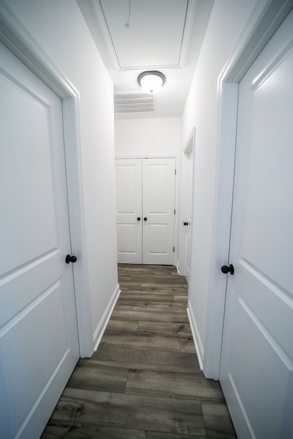 Hallway with light wood flooring, white baseboards, multiple white paneled doors featuring black knobs, and a section of black and white tile floor visible near one doorway.