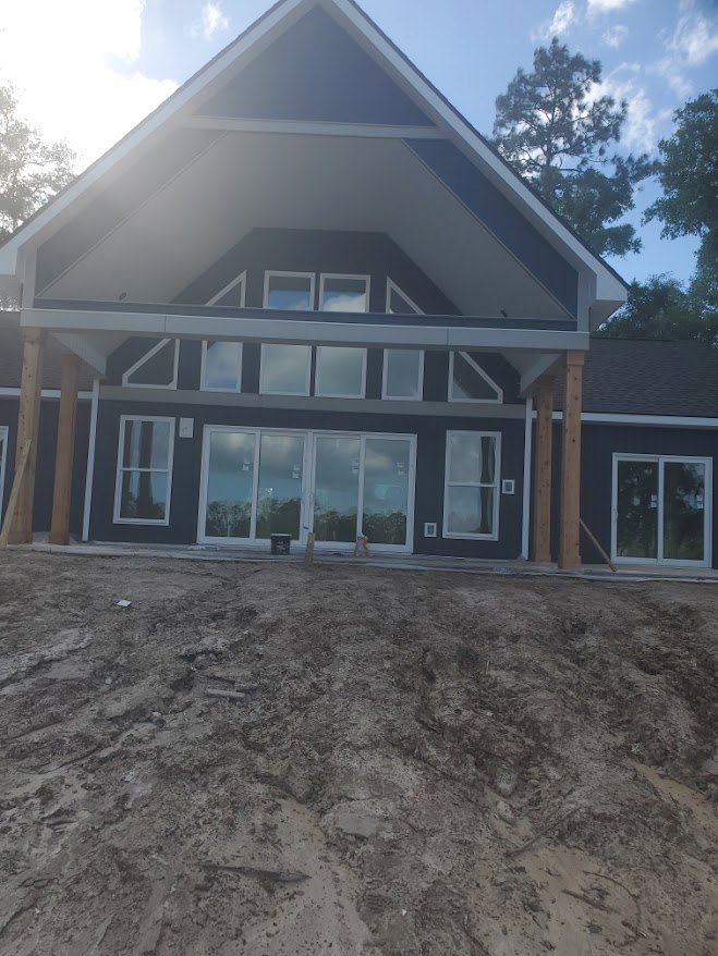 Partially built house with exposed wooden framing, multiple white-trimmed windows, triangular roof, dirt yard, and mature trees in the background under a blue sky with clouds