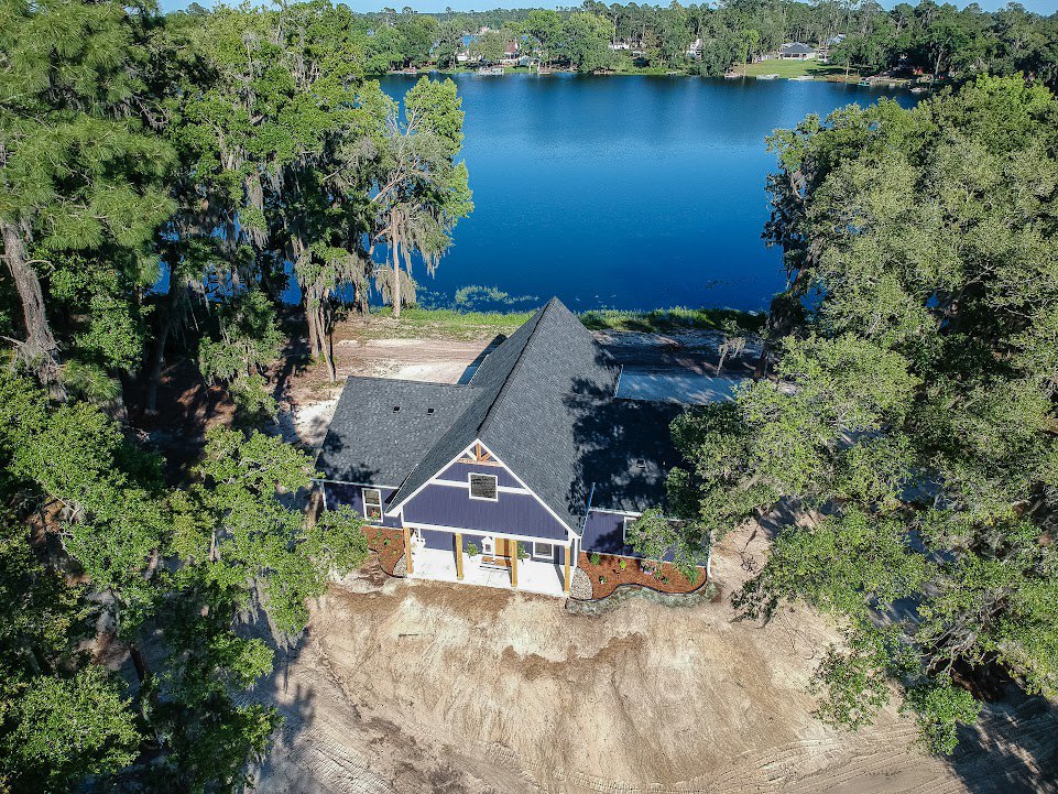 Two-story blue house with large roof and wraparound porch nestled among tall leafy trees, overlooking a calm lake
