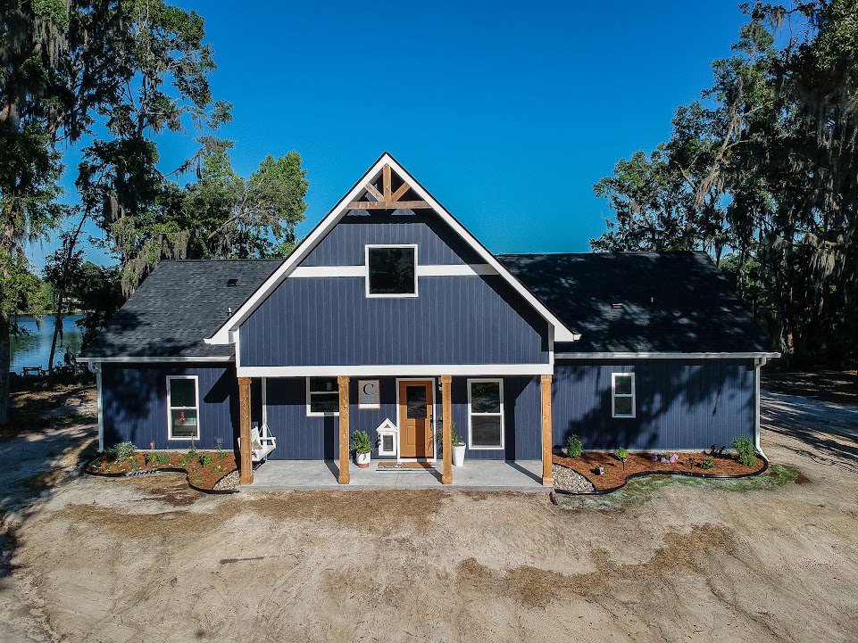 Blue-roofed house with white-framed windows, dirt driveway and patch in front, person working on roof, surrounded by trees and open sky