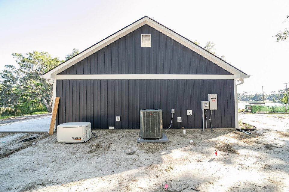 Grey stucco exterior wall with two mounted air conditioning units, small shed, waste container, and surrounding trees on landscaped property