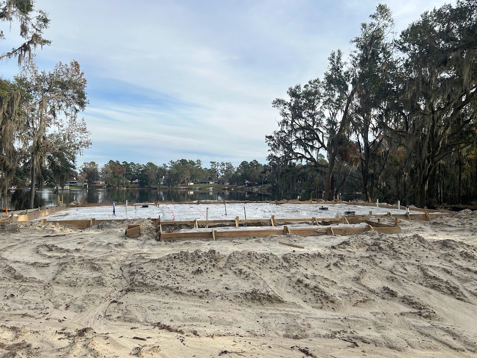 Sandy lakeshore bordered by tall trees with mossy branches, wooden frame structure near water, blue sky with scattered clouds