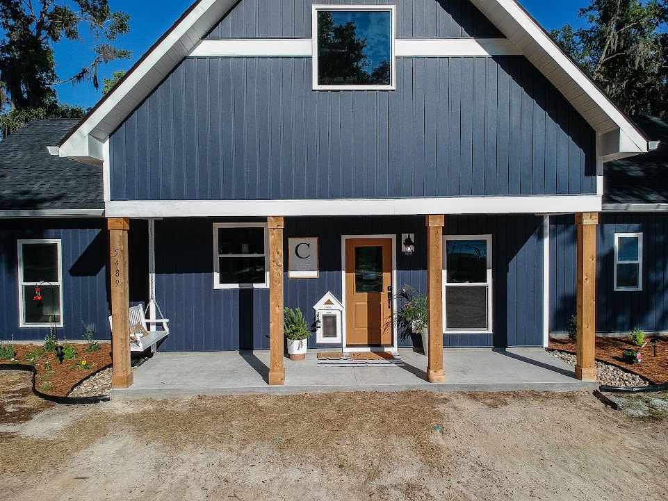 Blue siding with white trim, front porch, white entry door with window, white sign displaying black letter, windows reflecting trees, red light bulb visible through window